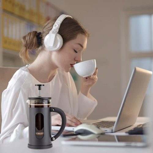 Woman drinking coffee and using a laptop with a French press on the table.